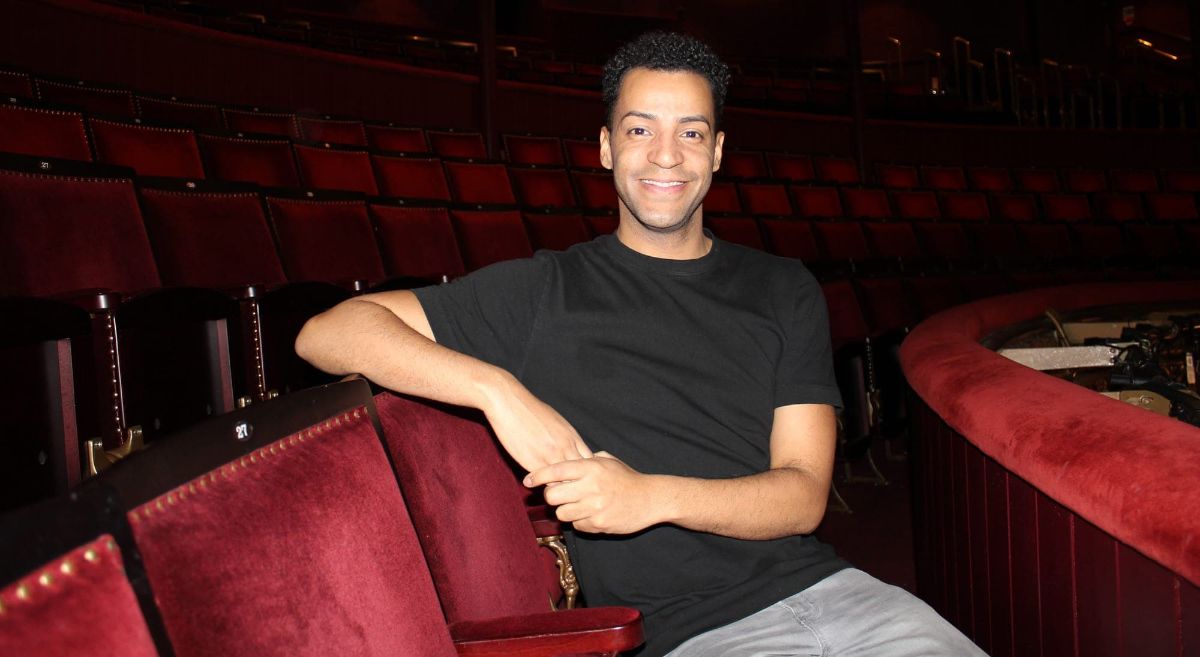 Male with dark hair wearing black short sleeved teeshirt seated alone in theatre with red plush seats in background.